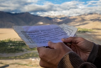 A person holds a piece of paper with text in an unidentified script, possibly Tibetan or Sanskrit, against a backdrop of expansive mountainous terrain under a partly cloudy sky.