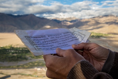 A person holds a piece of paper with text in an unidentified script, possibly Tibetan or Sanskrit, against a backdrop of expansive mountainous terrain under a partly cloudy sky.