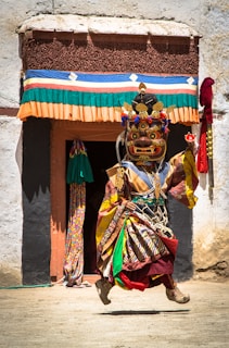 A person dressed in an elaborate and colorful traditional costume is performing a dance. The costume includes a large, ornate mask with intricate designs and bright colors. Behind them is a building with a doorway adorned with multicolored fabric. The scene is bright, suggesting daylight, with shadows cast on the ground.