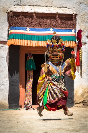 A person dressed in an elaborate and colorful traditional costume is performing a dance. The costume includes a large, ornate mask with intricate designs and bright colors. Behind them is a building with a doorway adorned with multicolored fabric. The scene is bright, suggesting daylight, with shadows cast on the ground.