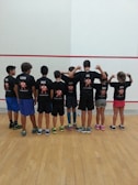 A group of children standing in a row facing away from the camera, wearing matching black t-shirts with colorful designs and names on the back. They are in a sport facility with a wooden floor and a white wall with red lines.