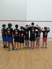 A group of children standing in a row facing away from the camera, wearing matching black t-shirts with colorful designs and names on the back. They are in a sport facility with a wooden floor and a white wall with red lines.