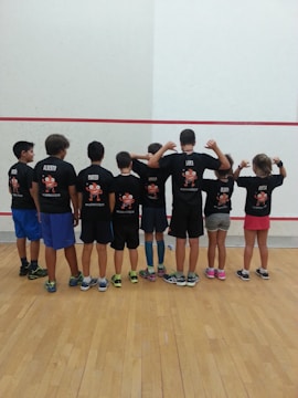 A group of children standing in a row facing away from the camera, wearing matching black t-shirts with colorful designs and names on the back. They are in a sport facility with a wooden floor and a white wall with red lines.