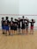 A group of children standing in a row facing away from the camera, wearing matching black t-shirts with colorful designs and names on the back. They are in a sport facility with a wooden floor and a white wall with red lines.