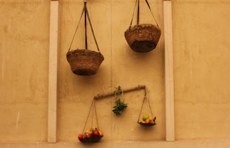 A rustic wall features traditional balance scales hanging vertically. The scales are made of rough, woven materials. One scale holds a variety of colorful fruits, while the other appears empty. The arrangement is simple, with a branch supporting a small plant and the balanced fruits.