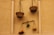A rustic wall features traditional balance scales hanging vertically. The scales are made of rough, woven materials. One scale holds a variety of colorful fruits, while the other appears empty. The arrangement is simple, with a branch supporting a small plant and the balanced fruits.
