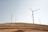 Wind turbines stand on a gently rolling, dry landscape under a clear sky. The turbines are evenly spaced across the field, casting long shadows on the light brown earth. The scene conveys a sense of open space and sustainable energy production.