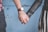 Two hands are interlocked, suggesting a gesture of affection or companionship. One person wears a leather jacket with a striped skirt, while the other is dressed in a plaid shirt and a watch on their wrist. The background consists of a textured blue wall.
