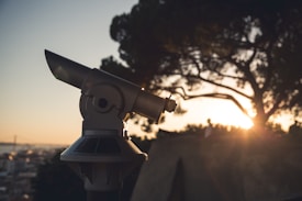 A telescope is positioned outdoors, focused towards a scenic view with the sun setting behind a silhouette of trees. The sky is clear, casting a warm glow over the landscape.