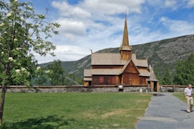A traditional wooden stave church with intricate designs stands against a backdrop of lush green hills and a partly cloudy sky. The church is surrounded by a stone wall and a well-maintained lawn. A paved path leads to the church entrance, and a tree with white blossoms is visible in the foreground. A person is walking along the path.