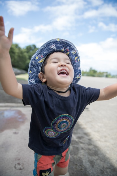 A joyful child with arms raised, wearing a colorful hat and a dark shirt with a circular pattern, standing outdoors on a sunny day with a clear blue sky.