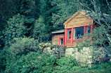 A rustic wooden cabin is situated amidst dense greenery, with a stone foundation and bright red-framed windows. The cabin is surrounded by lush trees and foliage, creating a serene and secluded atmosphere.