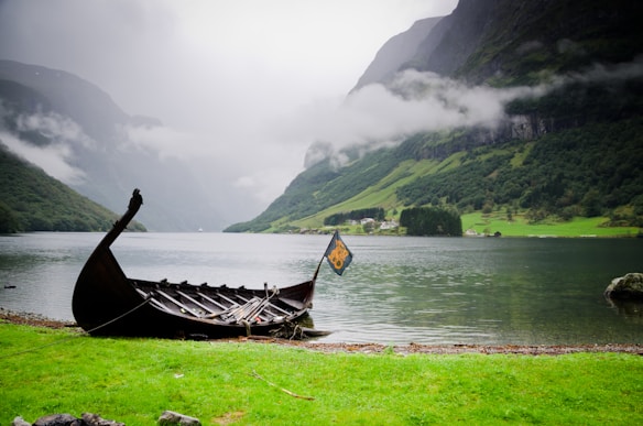 A traditional wooden Viking ship is docked by the edge of a serene fjord, surrounded by lush green hills and mist-covered mountains. The water is calm, reflecting the overcast sky and the surrounding landscape. A small flag attached to the ship flutters gently in the breeze.