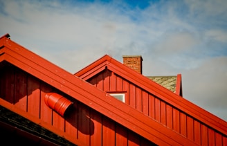 Red wooden rooftops with a chimney. The roof structure is sharply angled and features a cylindrical vent. The sky above is partly cloudy with patches of blue.