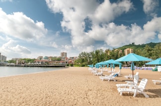 A sandy beach with a row of white lounge chairs and turquoise umbrellas. The background features a town with a coastline lined with buildings and green hills. The sky is filled with fluffy clouds, creating a bright and summery atmosphere.
