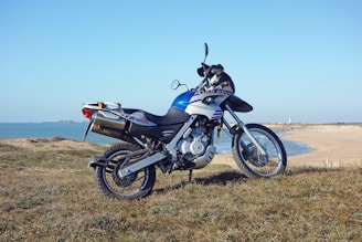 A motorcycle with a sleek design is parked on a grassy area overlooking a coastal landscape. The bike appears modern and sporty, featuring a prominent blue and white color scheme. In the background, a sandy beach and calm sea create a peaceful, open vista.