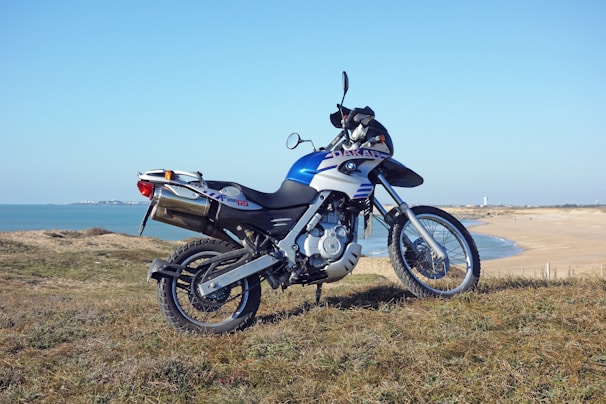 A motorcycle with a sleek design is parked on a grassy area overlooking a coastal landscape. The bike appears modern and sporty, featuring a prominent blue and white color scheme. In the background, a sandy beach and calm sea create a peaceful, open vista.
