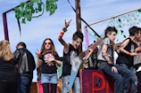 A group of young people dressed in punk rock attire are on a float decorated with green shamrocks. One person is holding a large, star-shaped guitar and making a rock-n-roll hand gesture. Another person sits to the side, appearing relaxed but also in similar punk attire. A young girl wearing sunglasses is making a hand gesture typically associated with rock music. There are onlookers in the background, and the scene seems lively, possibly part of a parade or festival.