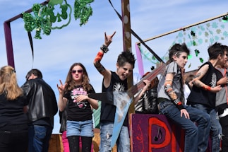 A group of young people dressed in punk rock attire are on a float decorated with green shamrocks. One person is holding a large, star-shaped guitar and making a rock-n-roll hand gesture. Another person sits to the side, appearing relaxed but also in similar punk attire. A young girl wearing sunglasses is making a hand gesture typically associated with rock music. There are onlookers in the background, and the scene seems lively, possibly part of a parade or festival.