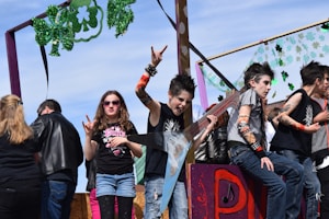 A group of young people dressed in punk rock attire are on a float decorated with green shamrocks. One person is holding a large, star-shaped guitar and making a rock-n-roll hand gesture. Another person sits to the side, appearing relaxed but also in similar punk attire. A young girl wearing sunglasses is making a hand gesture typically associated with rock music. There are onlookers in the background, and the scene seems lively, possibly part of a parade or festival.