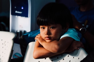 A young child with a thoughtful expression rests their chin on their arms, which are placed on the back of a white perforated plastic chair. The lighting casts a gentle shadow across the child's face, highlighting their contemplative mood. The background is dimly lit, with hints of a blue water dispenser and another person partially visible, adding depth to the scene.