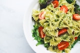 A white bowl filled with bow-tie pasta coated in a vibrant green pesto sauce, garnished with fresh cherry tomato halves and nestled on a bed of mixed greens.