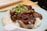 A dish of roasted duck served on a bed of white rice, garnished with a fresh green onion and ginger topping. The food is presented on a light blue plate with a white soup spoon on the side. Wooden chopsticks are placed in the background.
