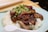 A dish of roasted duck served on a bed of white rice, garnished with a fresh green onion and ginger topping. The food is presented on a light blue plate with a white soup spoon on the side. Wooden chopsticks are placed in the background.