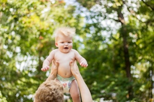 A baby with light hair and wearing a diaper is being lifted into the air by a pair of adult hands. The background is a lush, out-of-focus green foliage, suggesting a natural outdoor setting.
