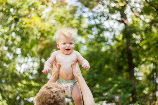 A baby with light hair and wearing a diaper is being lifted into the air by a pair of adult hands. The background is a lush, out-of-focus green foliage, suggesting a natural outdoor setting.