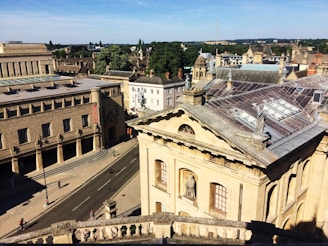 Aerial view of a historic cityscape featuring classical architecture with ornate facades and statues on the rooftops. The street below is lined with bicycles and a few pedestrians. Trees and additional buildings are visible in the background under a clear blue sky.