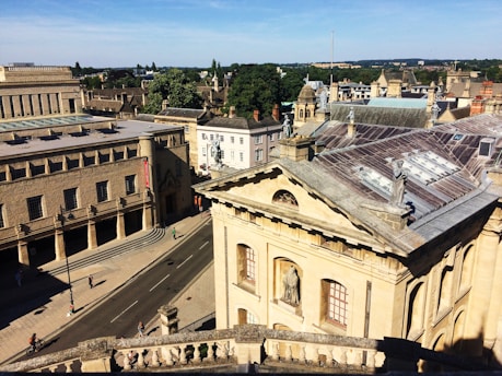 Aerial view of a historic cityscape featuring classical architecture with ornate facades and statues on the rooftops. The street below is lined with bicycles and a few pedestrians. Trees and additional buildings are visible in the background under a clear blue sky.