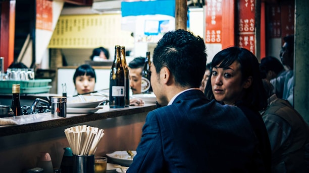 A group of people sits at a sushi bar with various dishes and bottles on the counter. In the foreground, a woman looks towards her companion, who is dressed in a suit, while others are seen engaging in conversation or watching the chef work behind the bar. The setting gives a cozy and busy atmosphere typical of a small Japanese eatery.