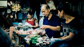 A bustling food stall with people preparing and serving a variety of skewered meats and vegetables. The setting appears to be a small, cozy eatery, filled with decorative items and a lively atmosphere. Two men are focused on food preparation while a woman in the background is engaged with wrapping or cleaning.