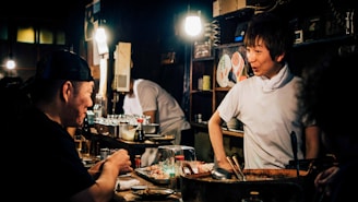 A cozy, dimly-lit restaurant scene featuring a chef interacting with a patron at the counter. The interior is rustic with warm lighting highlighting the chef's friendly expression. Various culinary tools and dishes are near the countertop, with shelves containing plates and cups in the background.