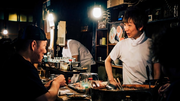 A cozy, dimly-lit restaurant scene featuring a chef interacting with a patron at the counter. The interior is rustic with warm lighting highlighting the chef's friendly expression. Various culinary tools and dishes are near the countertop, with shelves containing plates and cups in the background.