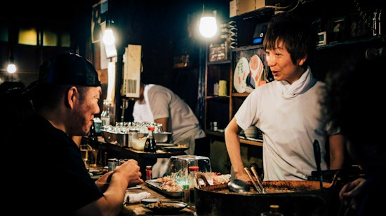 A cozy, dimly-lit restaurant scene featuring a chef interacting with a patron at the counter. The interior is rustic with warm lighting highlighting the chef's friendly expression. Various culinary tools and dishes are near the countertop, with shelves containing plates and cups in the background.