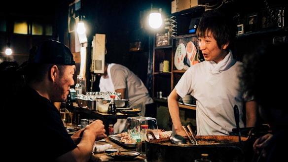 A cozy, dimly-lit restaurant scene featuring a chef interacting with a patron at the counter. The interior is rustic with warm lighting highlighting the chef's friendly expression. Various culinary tools and dishes are near the countertop, with shelves containing plates and cups in the background.