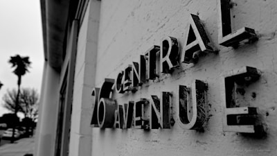 A black and white photograph featuring a close-up view of vintage-style 3D letters mounted on a textured wall, spelling '26 Central Avenue'. The perspective is angled, with focus on the lettering's aged and worn texture. In the background, silhouettes of palm trees and buildings are slightly blurred, contributing to the image's depth.