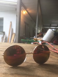 Round, reflective sunglasses are placed on a wooden surface in a workshop or garage-like setting. The reflection in the lenses shows partial features of a person. The background includes various workshop materials, such as buckets, cables, and wooden planks, with an overall dim lighting from a hanging bulb.