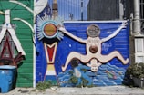 A vibrant wall mural features a stylized human figure with outstretched limbs against a bright blue background. The figure is adorned with circular patterns and is surrounded by abstract shapes, including a sun-like element. The left side of the mural has a green wooden panel with decorative elements and a blue recycling bin in the foreground. Plants grow at the base of the mural, adding a touch of greenery.