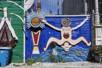 A vibrant wall mural features a stylized human figure with outstretched limbs against a bright blue background. The figure is adorned with circular patterns and is surrounded by abstract shapes, including a sun-like element. The left side of the mural has a green wooden panel with decorative elements and a blue recycling bin in the foreground. Plants grow at the base of the mural, adding a touch of greenery.