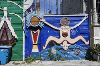 A vibrant wall mural features a stylized human figure with outstretched limbs against a bright blue background. The figure is adorned with circular patterns and is surrounded by abstract shapes, including a sun-like element. The left side of the mural has a green wooden panel with decorative elements and a blue recycling bin in the foreground. Plants grow at the base of the mural, adding a touch of greenery.