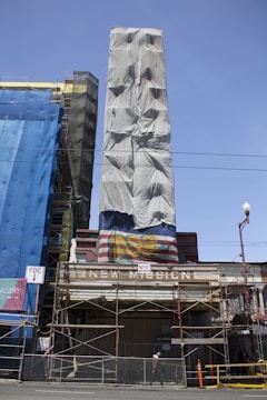 A partially covered building facade with scaffolding in front. The covering has a graffiti image of the word 'BOP' in vibrant colors and sections of cloth are draped down. Next to it is another building also wrapped in a blue cloth, with signs and construction barriers visible. The sky is clear and blue.