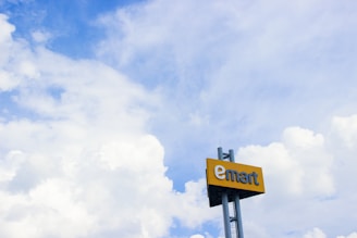 A large sign with the word 'emart' is mounted on a pole against a backdrop of a bright blue sky filled with fluffy white clouds.