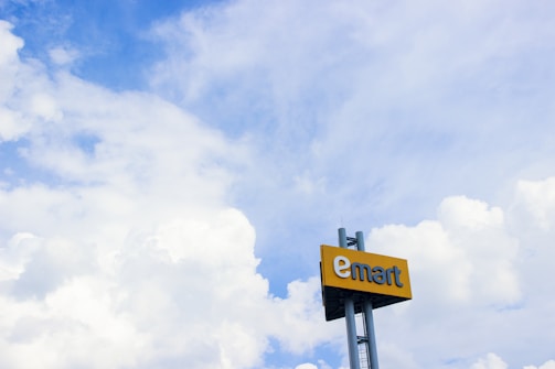 A large sign with the word 'emart' is mounted on a pole against a backdrop of a bright blue sky filled with fluffy white clouds.