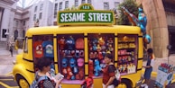 A yellow vehicle resembling a bus is adorned with a 'Sesame Street' sign. The vehicle is converted into a shop displaying a variety of merchandise including plush toys of characters like Elmo, Cookie Monster, and others. Several people are standing around the bus, some browsing items. The setting appears to be an urban environment with buildings in the background.