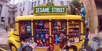 A yellow vehicle resembling a bus is adorned with a 'Sesame Street' sign. The vehicle is converted into a shop displaying a variety of merchandise including plush toys of characters like Elmo, Cookie Monster, and others. Several people are standing around the bus, some browsing items. The setting appears to be an urban environment with buildings in the background.