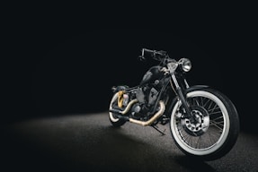 A sleek black motorcycle with a matte finish, displayed against a dark background. The bike features whitewall tires, chrome accents, and a low seat design, creating a classic yet modern look. The headlight is prominently positioned, and the handlebar is low, enhancing its streamlined appearance.