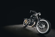A sleek black motorcycle with a matte finish, displayed against a dark background. The bike features whitewall tires, chrome accents, and a low seat design, creating a classic yet modern look. The headlight is prominently positioned, and the handlebar is low, enhancing its streamlined appearance.